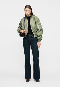 Νεαρή γυναίκα με πράσινο μπουφάν τύπου bomber, μαύρο τοπ, σκούρο μπλε τζιν, καφέ παπούτσια και κρατώντας μια μικρή σκούρα καφέ τσάντα ώμου.