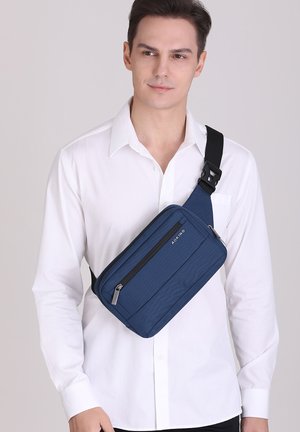 Sac bandoulière bleu marine en tissu texturé, doté d'un compartiment zippé, d'une sangle noire réglable et d'un design minimaliste.