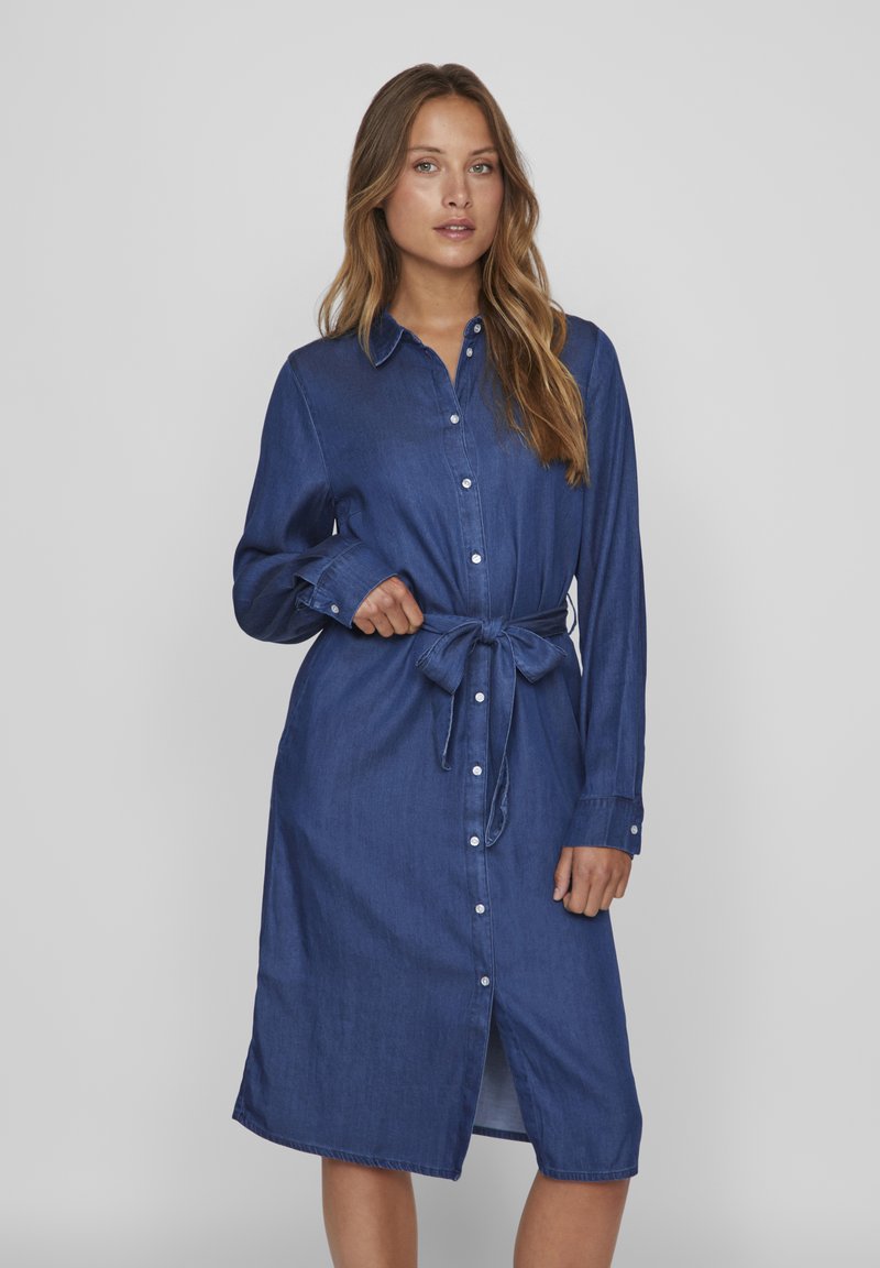 Denim srajčna obleka, dolgi rokavi, gumbnica spredaj, pas z usnjem, temno modra barva, dolžina do kolen, mehak občutek, subtilen lesk, brez vzorcev.