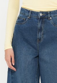 Jeans de mezclilla azul con cintura alta, dos bolsillos frontales y un solo botón metálico de cierre. La mezclilla tiene una textura sutil y un lavado medio.