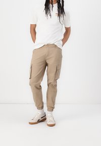 Beige Cargohose mit Seitentaschen, verjüngten Bündchen und Knopfverschluss. Kombiniert mit einem weißen T-Shirt und weißen Sneakern mit schwarzen Details.