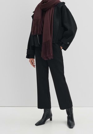 Persoon met zwarte cropped broek, zwarte laarzen met hak, een oversized zwart jasje en een lange donker bordeauxrode sjaal tegen een effen achtergrond.