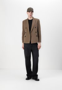 Blazer marrón de doble botonadura con botones negros, usado sobre una camisa negra. Combinado con jeans de pierna ancha oscuros y zapatos negros. Incluye una gorra de cuadro.