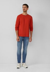 Rotes Langarm-T-Shirt, weicher Baumwollstoff, Rundhalsausschnitt; kombiniert mit hellblauen Skinny-Jeans und beigen Sportschuhen.