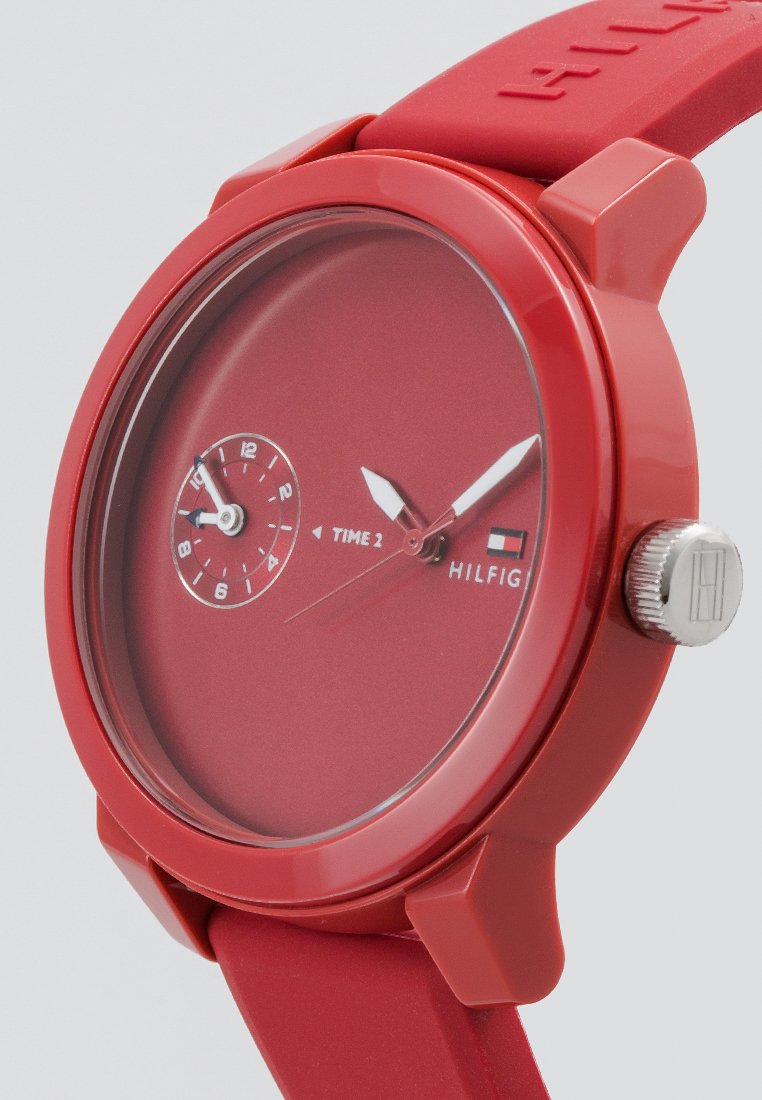 Reloj de pulsera rojo con dos esferas de tiempo, agujas plateadas y correa de silicona roja, que muestra el nombre y el logotipo de la marca en la esfera.