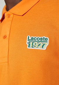 Gros plan sur un polo orange avec le logo Lacoste 1927 vert et jaune sur la poitrine gauche près des boutons.