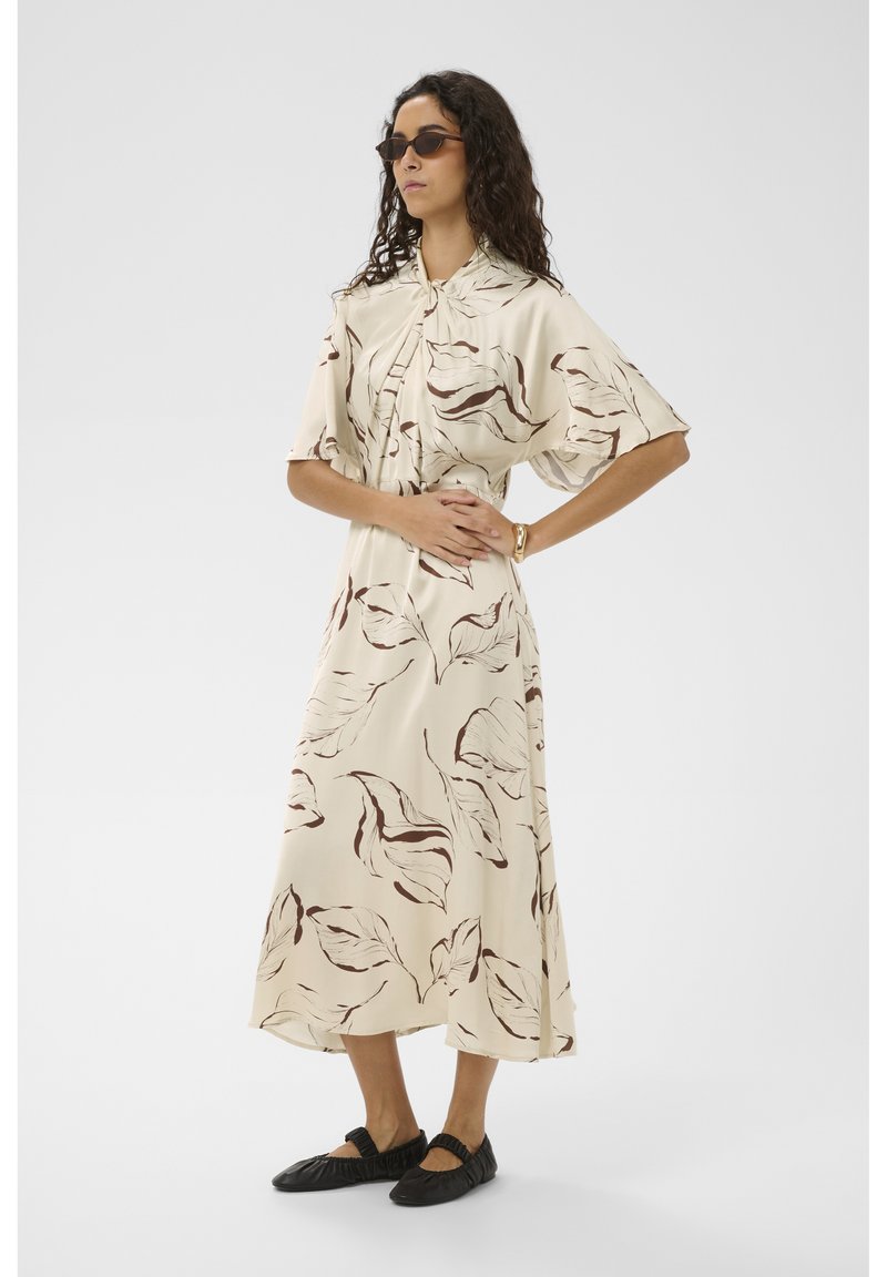 Vrouw met lang krullend haar, een zonnebril, een beige jurk met bruine bladmotieven, zwarte ballerina's en een gouden armband, staand met gekruiste armen.