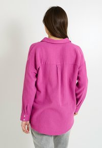 Camisa de manga larga fucsia con cuello, tejido texturizado y puños abotonados. Presenta un pliegue en la parte posterior y un dobladillo curvado para un ajuste relajado.