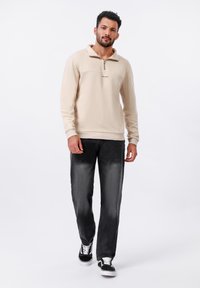Pull à col zippé beige avec un panneau horizontal contrastant, associé à un jean noir délavé et des sneakers noires avec des accents blancs.