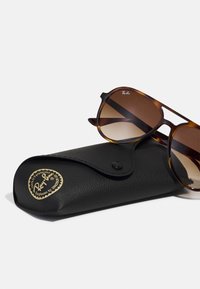Occhiali da sole Ray-Ban con lenti marroni sfumate in una montatura tartaruga, adagiati su un astuccio in pelle nera con dettagli del logo in oro.