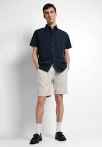 Mörkblå kortärmad skjorta med knapplist, tillsammans med krämfärgade shorts. Modellen har svarta skor med vita strumpor och står med händerna i fickorna.