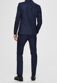 Mężczyzna w dopasowanym granatowym garniturze i czarnych eleganckich butach, stojący z jedną ręką w kieszeni, odwrócony plecami do aparatu.