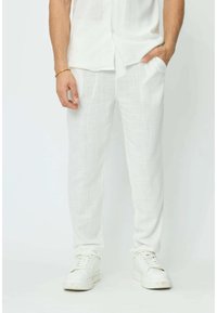 Pantalons en lin blancs avec une texture légère, offrant une coupe décontractée, des poches latérales et un motif tissé subtil. Associés à des baskets blanches.
