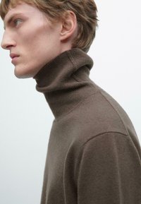 Brązowy sweter typu golf wyk wykonany z miękkiego materiału, z żeberkowym wykończeniem kołnierza i gładką teksturą, pokazany noszony z boku.