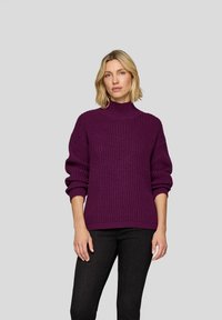 Gestickter Pullover in tiefem Lila, mit hohem Kragen, gerippter Textur und abfallenden Schultern. Kombiniert mit dunklen Jeans, vor einem schlichten Hintergrund stehend.