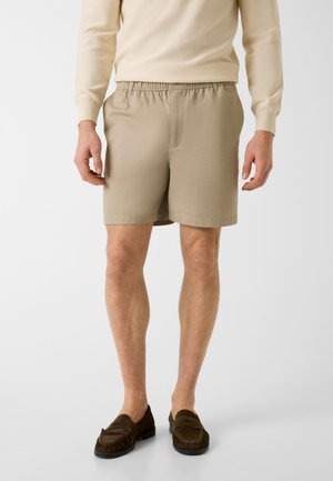 Homme portant un short beige à taille élastique, une chemise crème à manches longues et des mocassins marron, debout devant un fond blanc uni.