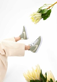 Gröna högre sneakers i canvas med vita accenter, ihopparade med ljusbeiga byxor, placerade ovanför gulaktigt vita blommor.