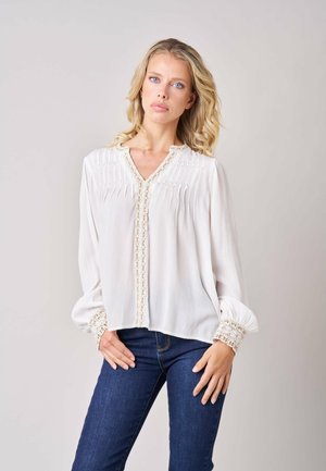 Femme blonde aux cheveux bouclés portant un chemisier blanc à manches longues avec bordure en dentelle et un jean bleu, debout devant un fond uni.