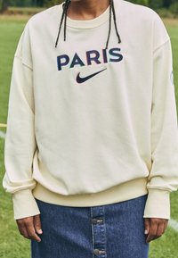 Persona con cabello trenzado que lleva un sudadero crema con "PARIS" y el logo de Nike, combinado con una falda de mezclilla abotonada, de pie sobre el césped.