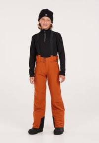 Pantaloni da neve arancioni con dettagli neri, bretelle regolabili e vita alta. Indossati con una maglia nera a maniche lunghe e un berretto nero.