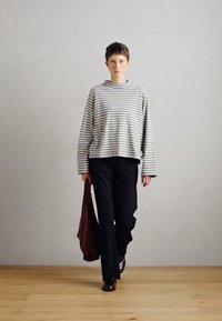 Camisola listrada de mangas longas em cinza e branco, combinada com calças largas pretas e uma bolsa bordeaux, em pé sobre um chão de madeira contra um fundo neutro.