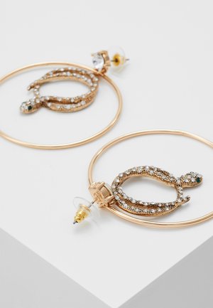 Gouden creolen met binnenin slangenpatronen bezet met strass, bevestigd met doorzichtige en gouden vlinderklemmen op een witte ondergrond.