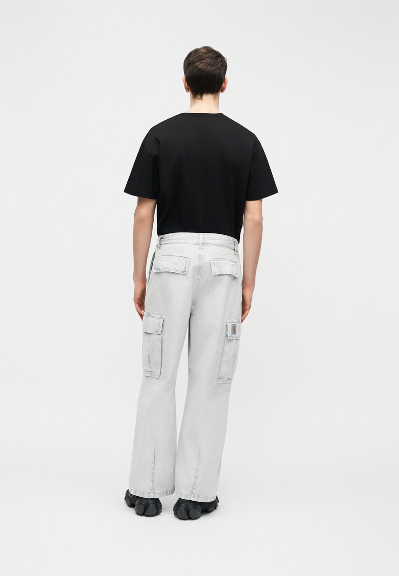 Person steht mit dem Rücken zur Kamera, trägt ein schwarzes kurzärmeliges Hemd, hellgraue Cargo-Hosen mit Klappentaschen und schwarze Schuhe.