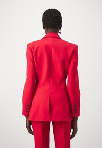 Červený blazer se strukturovanými rameny, s jedním rozventilovaným zadním dílem a čtyřmi knoflíkovými detaily na rukávech. Hladká tkanina s dokonale padnoucím střihem, doplněná o odpovídající kalhoty.