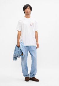 Λευκό βαμβακερό T-shirt με μπροστινό γραφικό σχέδιο, ανοιχτό μπλε τζιν παντελόνι και μπλε τζιν μπουφάν κρατημένο στο χέρι. Καφέ παπούτσια ολοκληρώνουν το σύνολο.