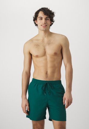 Bañador verde con cinturilla elástica y cordón, textura suave, largo por encima de la rodilla, con un diseño minimalista.