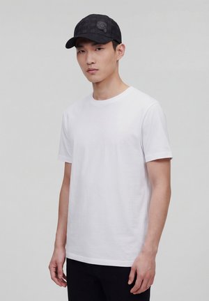 Młody mężczyzna w gładkiej białej koszulce z krótkim rękawem i czarnej czapce z daszkiem z wzorem na jasnoszarym tle.