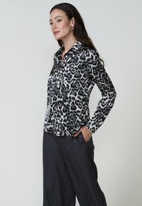 Il modello sta in piedi con sicurezza, indossando una camicia con stampa leopardata bianca e nera abbinata a pantaloni scuri, con le mani in tasca, su uno sfondo chiaro.