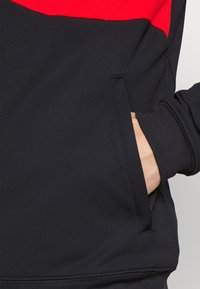 Sudadera negra con una franja roja horizontal en la parte superior, con puños de canalé y un bolsillo lateral. Textura suave, diseño casual.