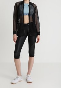 Woman wearing black mesh zip-up jacket, light blue sports bra, black cropped leggings with mesh panels, and white athletic shoes.