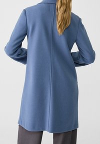 Dlouhý modrý kabát s jemnou texturou, jediným zadním švem, širokými klopami a volným střihem, doplněný o šedé kalhoty.