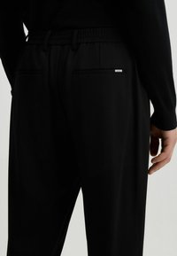 Schwarze Hosen mit elastischem Bund, zwei Gesäßtaschen und einem kleinen Metallelement auf der rechten Tasche. Glatte Stoffoberfläche.
