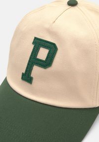 Căciulă bej cu vizieră verde și un "P" brodat verde în față. Prezintă un material structurat și o coroană rotunjită.