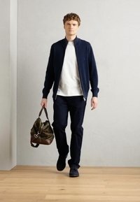 Chaqueta de cremallera azul marino sobre una camiseta blanca, combinada con pantalones oscuros. Sostiene un bolso de color verde oliva con detalles marrones. Fondo de suelo de madera.