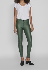 Pantalones skinnies de talle alto en verde, fabricados con un material brillante, que cuentan con un botón y cremallera en la parte delantera. Combinados con una camiseta blanca de cuello en V y tacones negros.