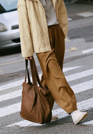 Henkilö kävelee suojatiellä yllään beige tikattu takki, ruskeat leveät housut, valkoiset lenkkarit ja suuri ruskea kauppakassi olalla.