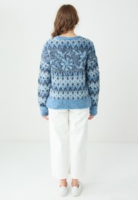 Gestrickter Pullover in verschiedenen Blautönen mit geometrischem Muster, Rundhalsausschnitt und geripptem Saum, kombiniert mit weißen Hosen mit weitem Bein und Turnschuhen.