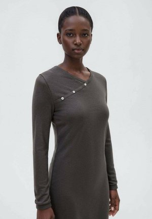 Donna con pelle scura e capelli raccolti indossa un vestito grigio a maniche lunghe con un dettaglio asimmetrico di bottoni vicino al collo.