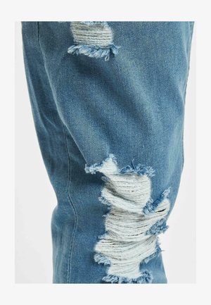 Sinised denim püksid, millel on kulunud plaastrid ja narmastega äärised, rõhutades kangast tekstuuri ja erinevaid siniseid toone.