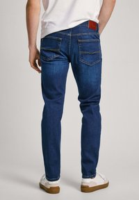 Jeans de mezclilla en azul oscuro con un corte entallado. Cuenta con bolsillos traseros con detalles de costura y una etiqueta de cuero marrón en la cintura.