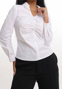 Camicia bianca a maniche lunghe con scollatura a V e dettaglio arricciato sul petto, dotata di polsini con bottoni e texture liscia, abbinata a pantaloni neri.