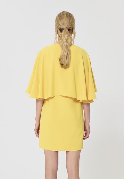 Femme avec une queue de cheval blonde portant une courte robe jaune avec un surcot ressemblant à une cape, debout de dos sur un fond blanc uni.