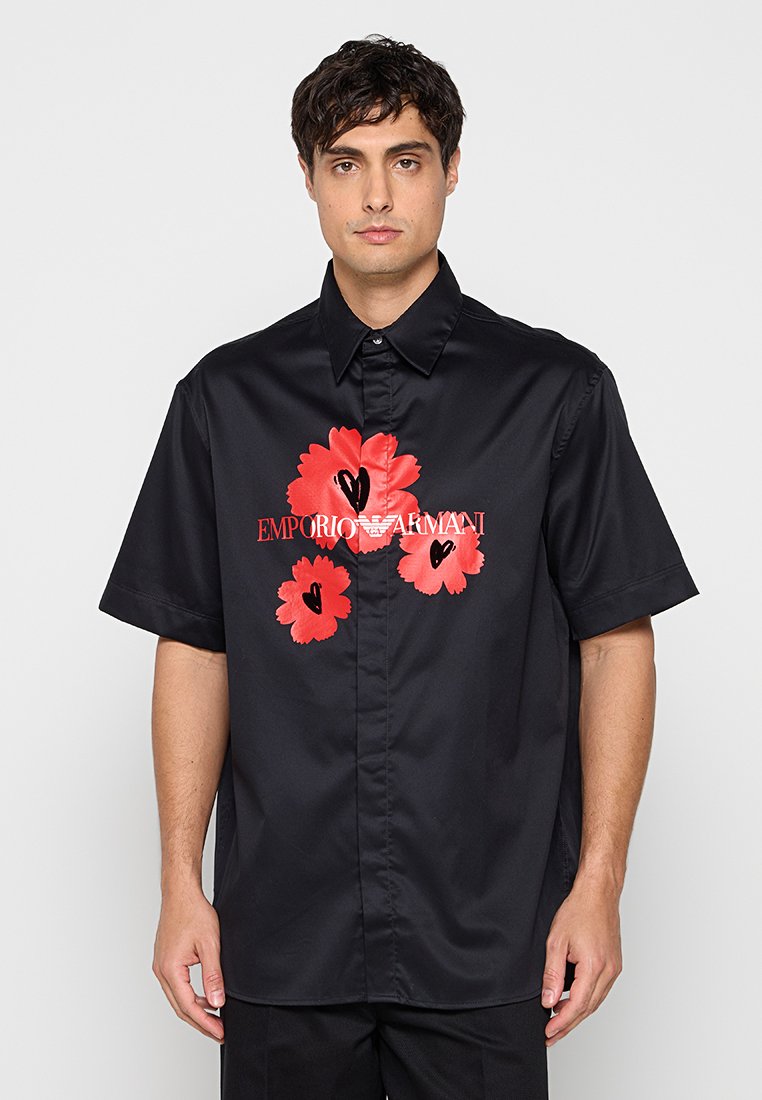Hombre con camisa negra de manga corta con estampado floral rojo y logo blanco de Emporio Armani en el frente, de pie contra un fondo gris.