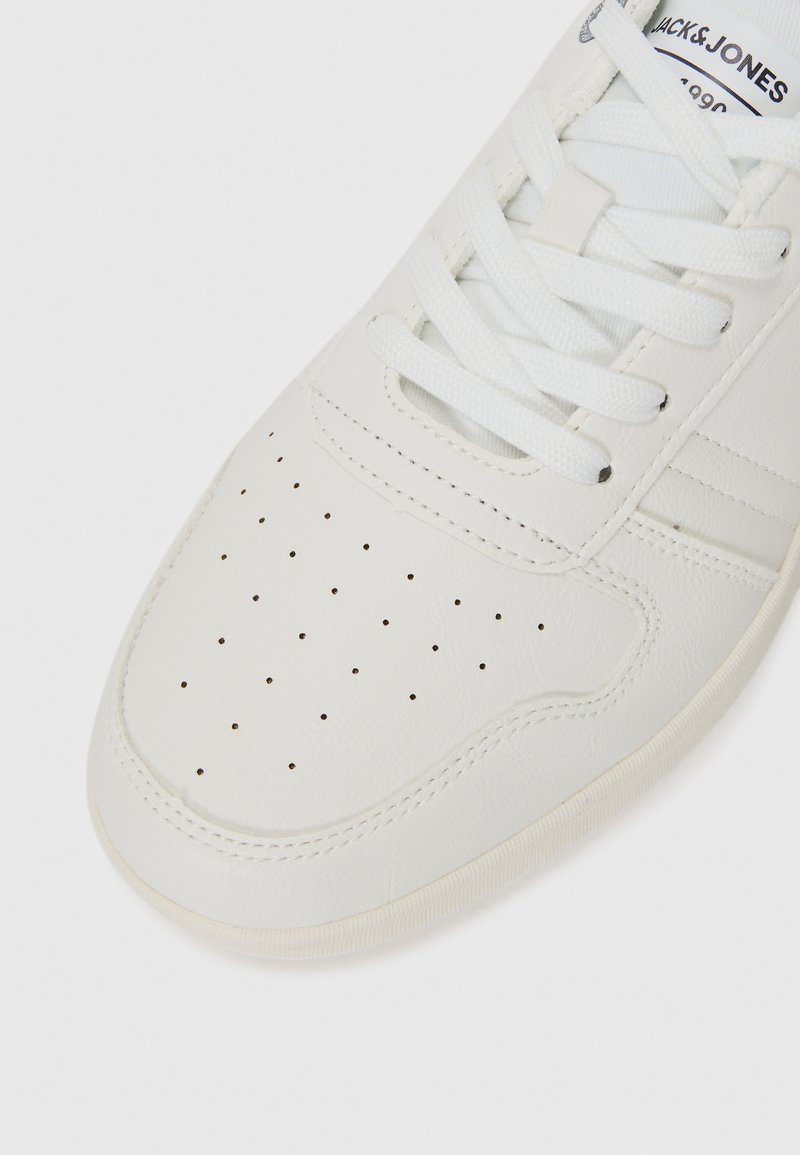 Gros plan sur une sneaker blanche présentant des perforations sur la boîte à orteils et des lacets blancs, avec un logo sur la languette. Design simple et minimaliste.