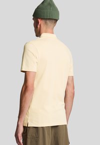 Lichtgele polo met korte mouwen, voorzien van een geribde kraag, zijsplitten en een textuurpatroon. Draagt met een olijfgroen mutsje.
