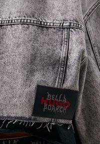 Farkkutakki haalistuneessa harmaassa, jossa on rispaantunut helma. Mukana on suorakulmainen musta etiketti, jossa lukee "BELLA HUGO" punaisella ja tyylitellyllä hopeanvärisellä kirjainmuodolla.